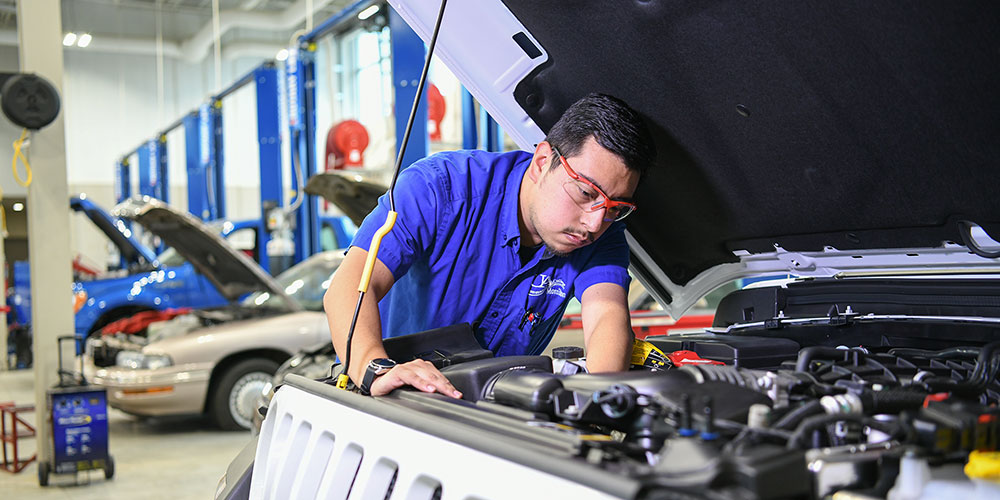 Student working under the hood of a car in the foreground in a shop with cars behind with their hoods up.