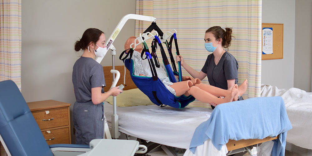 Two students in scrubs and mask learning to use the patient lift to move a mannequin from bed to chair.
