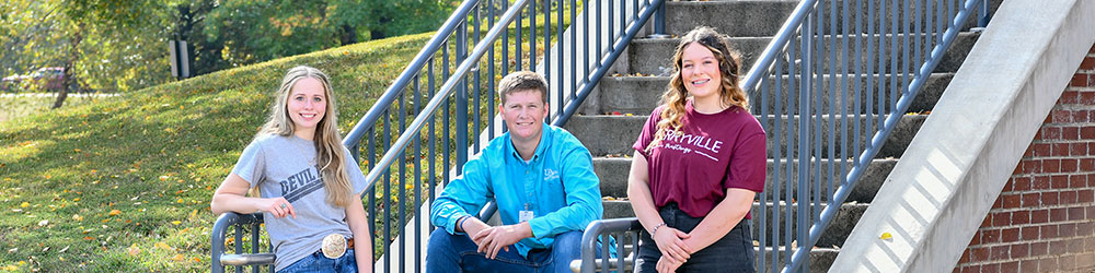 Three students from three different high schools posing at the base of an outside staircase, smiling for camera.